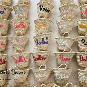 May include: A collection of small, straw tote bags with various names embroidered on them in different colors. The bags are arranged in rows on a white surface. The text "Marrakesh Decors" and "Handmade with Love" is visible at the bottom of the image.