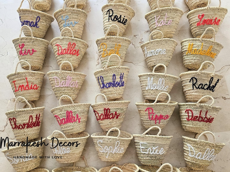 May include: A collection of small, straw tote bags with various names embroidered on them in different colors. The bags are arranged in rows on a white surface. The text "Marrakesh Decors" and "Handmade with Love" is visible at the bottom of the image.