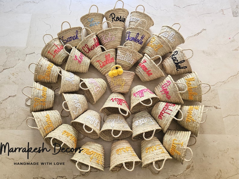 May include: A collection of 20 small, straw tote bags with personalized embroidered names in various colors. The bags are arranged in a circle on a light beige tiled surface. The text "Marrakesh Decor's" and "Handmade with Love" is visible at the bottom of the image.