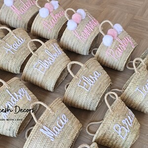 May include: A collection of straw tote bags with pom poms and personalized embroidery. The bags are in various shades of beige and have white embroidery with names like "Heidi", "Aubrey", "Kaisley", and "Elisa".