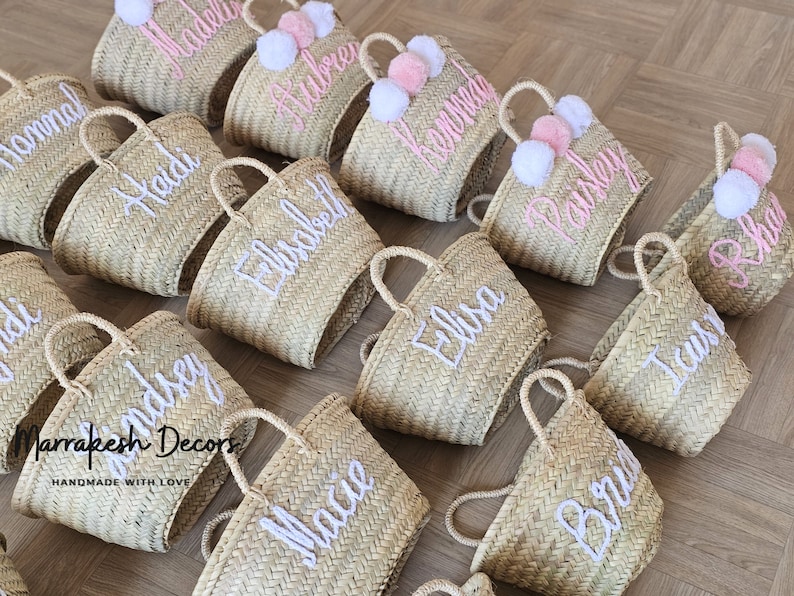May include: A collection of straw tote bags with pom poms and personalized embroidery. The bags are in various shades of beige and have white embroidery with names like "Heidi", "Aubrey", "Kaisley", and "Elisa".