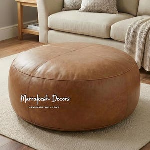Könnte beinhalten: Ein runder, brauner Lederhocker mit Steppdetails. Der Hocker steht auf einem hellen Teppich, mit einem beigen Sofa und einem hölzernen Beistelltisch im Hintergrund. Der Text "Marrakesh Decors HANDMADE WITH LOVE" ist auf dem Hocker aufgedruckt.