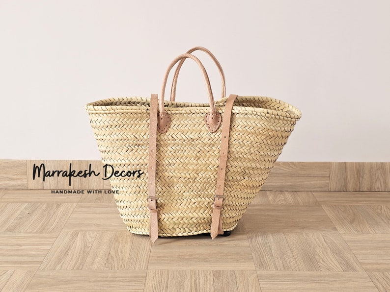 May include: A large, natural woven straw tote bag with brown leather straps and buckles. The bag is sitting on a wood floor. The text "Marrakesh Decors Handmade with Love" is visible at the bottom of the image.