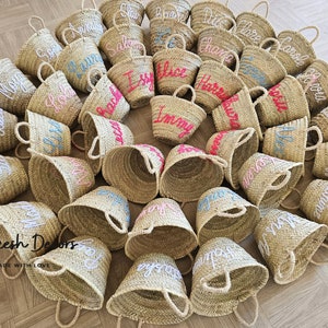May include: A collection of small, straw baskets with handles. Each basket is personalized with a name embroidered in white, pink, or blue thread. The baskets are arranged in a circle on a wooden floor. The text "Marrakesh Delors Handmade with Love" is visible in the bottom left corner of the image.