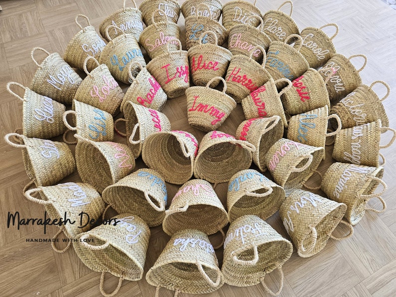 May include: A collection of small, straw baskets with handles. Each basket is personalized with a name embroidered in white, pink, or blue thread. The baskets are arranged in a circle on a wooden floor. The text "Marrakesh Delors Handmade with Love" is visible in the bottom left corner of the image.