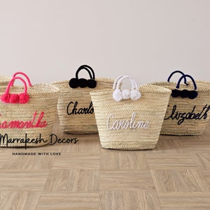 May include: Five straw tote bags with personalized names in colorful embroidery. The bags have pom poms in pink, white, and black. The bags are handmade with love by Marrakech Decors.