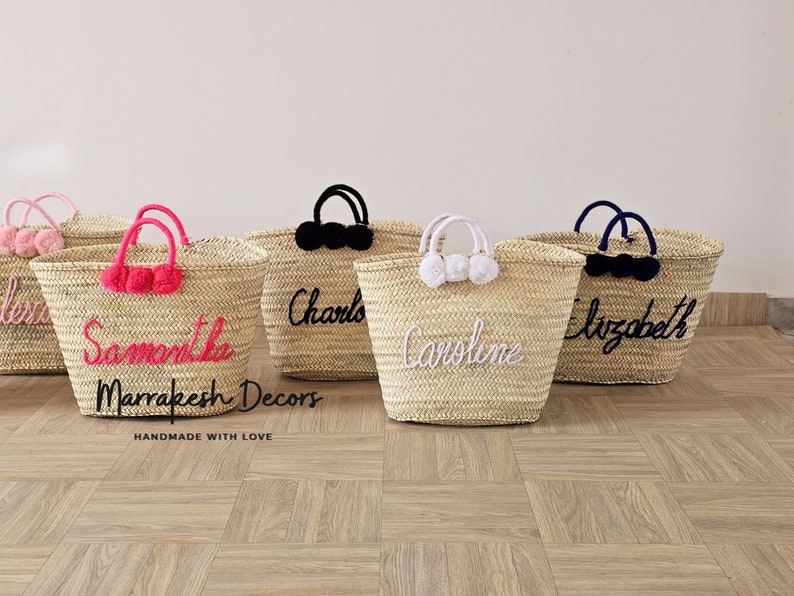 May include: Five straw tote bags with personalized names in colorful embroidery. The bags have pom poms in pink, white, and black. The bags are handmade with love by Marrakech Decors.