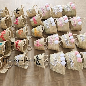 May include: A collection of small, natural woven baskets with personalized embroidery and pom-poms. The baskets are in various colors, including pink, orange, yellow, and green. The text "Marrakesh Decors Handmade with Love" is visible in the image.