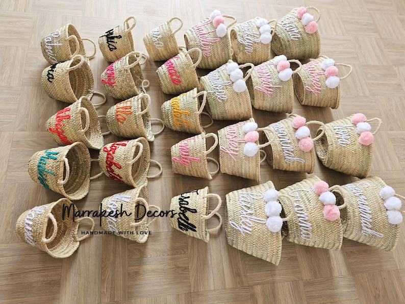 May include: A collection of small, natural woven baskets with personalized embroidery and pom-poms. The baskets are in various colors, including pink, orange, yellow, and green. The text "Marrakesh Decors Handmade with Love" is visible in the image.