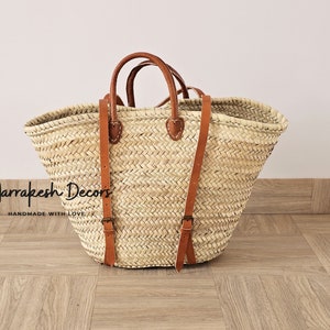 May include: A large, natural woven straw tote bag with brown leather straps and buckles. The bag has a wide, open top and is perfect for carrying groceries, books, or other items. The bag is handmade and has a rustic, natural look.