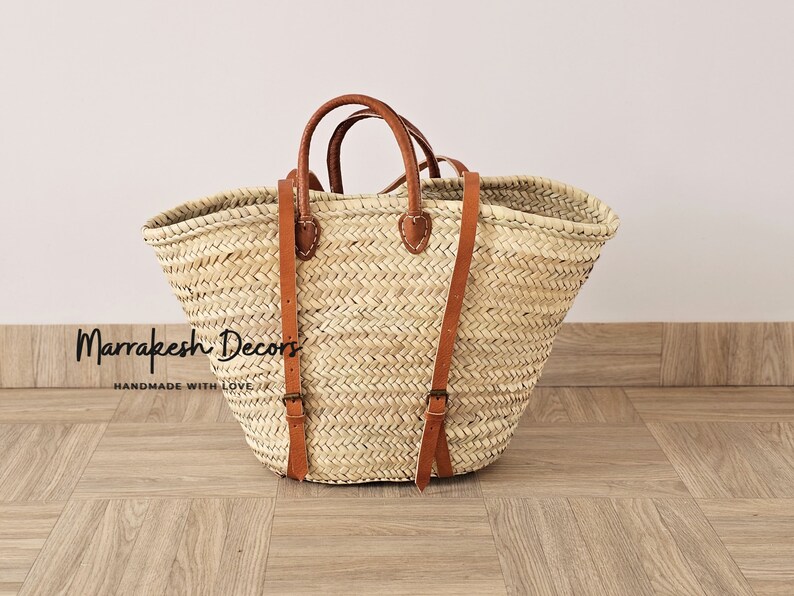 May include: A large, natural woven straw tote bag with brown leather straps and buckles. The bag has a wide, open top and is perfect for carrying groceries, books, or other items. The bag is handmade and has a rustic, natural look.