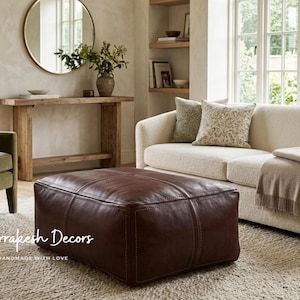 Könnte beinhalten: Ein großer, quadratischer Sitzhocker aus dunkelbraunem Leder mit sichtbaren Nähten an der Oberseite und den Seiten. Der Text Marrakesh Decors HANDMADE WITH LOVE ist in Weiß in der unteren linken Ecke des Hockers aufgedruckt.