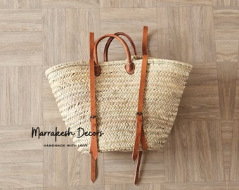 May include: A large, woven straw tote bag with brown leather straps and buckles. The bag is on a wood floor. The text "Marrakesh Decors Handmade with Love" is printed on the bag.