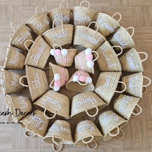 May include: A collection of 20 small, natural woven straw tote bags with white embroidered names. The bags are arranged in a circle on a wood floor. The bags have white pom poms attached to the handles.
