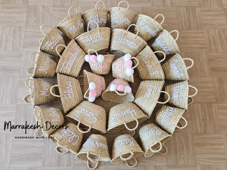 May include: A collection of 20 small, natural woven straw tote bags with white embroidered names. The bags are arranged in a circle on a wood floor. The bags have white pom poms attached to the handles.
