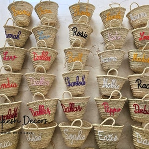 May include: A collection of small, straw tote bags with various names embroidered on them in different colors. The bags are arranged in rows on a white surface. The text "Marrakesh Decors" and "HANDMADE WITH LOVE" is visible at the bottom of the image.