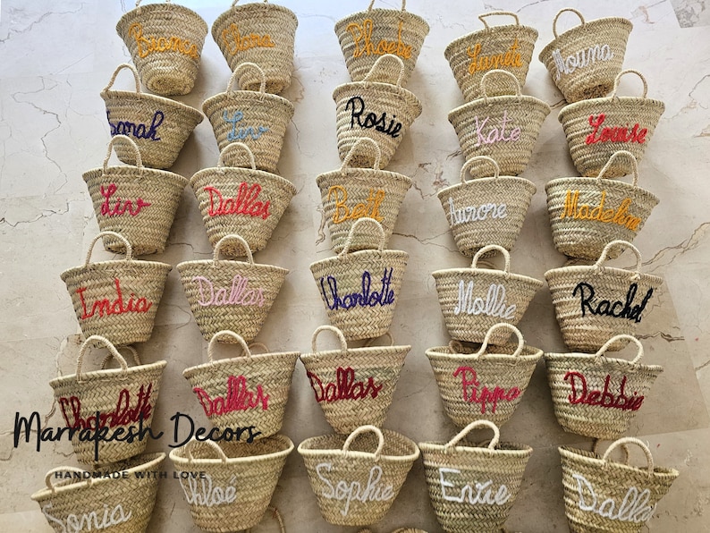 May include: A collection of small, straw tote bags with various names embroidered on them in different colors. The bags are arranged in rows on a white surface. The text "Marrakesh Decors" and "HANDMADE WITH LOVE" is visible at the bottom of the image.