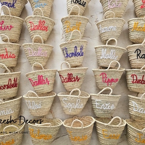 May include: A collection of small, woven straw bags with various names embroidered on them in different colors. The bags are arranged in rows on a light-colored surface. The text "Marrakesh Decors" and "HANDMADE WITH LOVE" is visible at the bottom of the image.
