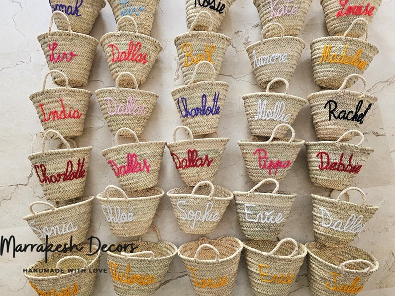 May include: A collection of small, woven straw bags with various names embroidered on them in different colors. The bags are arranged in rows on a light-colored surface. The text "Marrakesh Decors" and "HANDMADE WITH LOVE" is visible at the bottom of the image.
