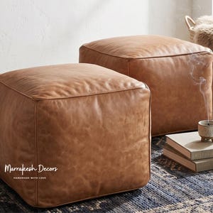 Könnte beinhalten: Zwei quadratische, braune Lederhocker. Die Hocker stehen auf einem blau-weiß gemusterten Teppich. Die Hocker haben ein Nahtdesign an den Kanten. Der Text "Marrakesh Decors Handmade with Love" ist auf der Vorderseite des Hockers zu sehen.
