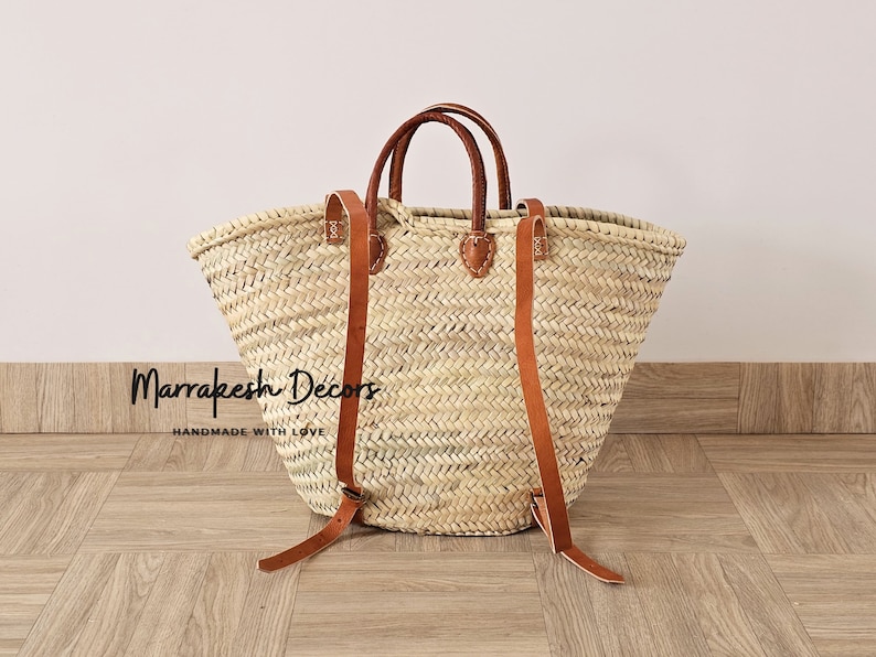 May include: A large, natural woven straw tote bag with brown leather handles and straps. The bag has a wide, open top and a rounded bottom. The straps are long enough to be worn as a backpack. The bag is sitting on a wooden floor. "Marrakesh Decors Handmade with Love" is printed on the bag.