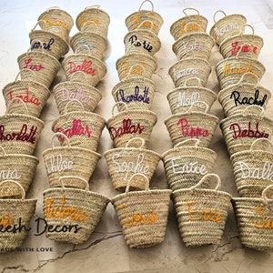 May include: A collection of straw tote bags with personalized names embroidered in various colors, including red, white, yellow, and pink. The bags are arranged in rows on a tiled floor. The text "Marrakesh Decors Handmade with Love" is visible at the bottom of the image.
