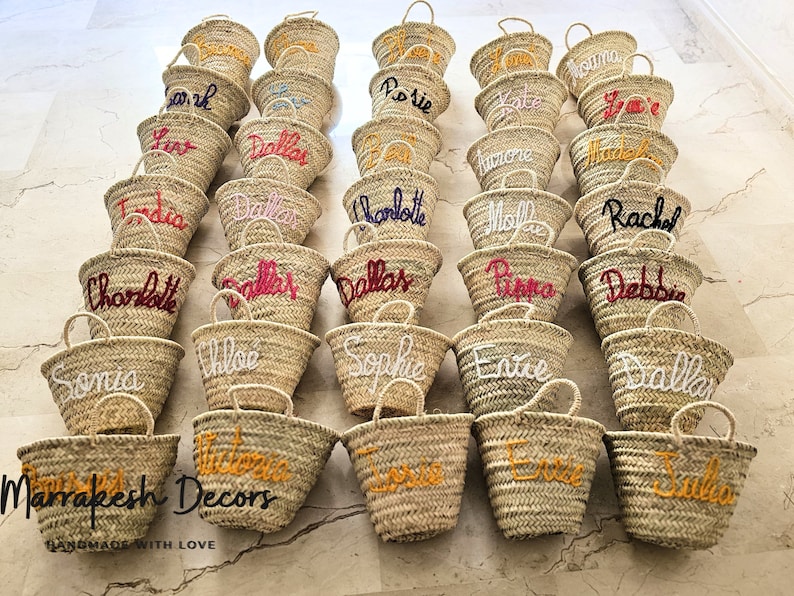 May include: A collection of straw tote bags with personalized names embroidered in various colors, including red, white, yellow, and pink. The bags are arranged in rows on a tiled floor. The text "Marrakesh Decors Handmade with Love" is visible at the bottom of the image.