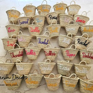 May include: A collection of straw tote bags with personalized names embroidered in various colors. The bags are arranged in a grid pattern on a light-colored surface. The text "Marrakesh Decors" and "HANDMADE WITH LOVE" is visible at the bottom of the image.