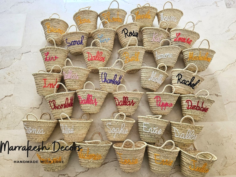 May include: A collection of straw tote bags with personalized names embroidered in various colors. The bags are arranged in a grid pattern on a light-colored surface. The text "Marrakesh Decors" and "HANDMADE WITH LOVE" is visible at the bottom of the image.
