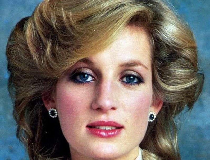 May include: A woman with blonde hair styled in a bouffant, wearing a white collared shirt and earrings. Her makeup features blue eyeshadow and a light pink lipstick.