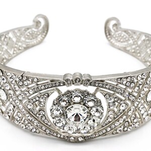 Puede incluir: Una diadema plateada con un dise&ntilde;o delicado e intrincado que presenta un gran grupo de diamantes de imitaci&oacute;n transparentes en el centro.