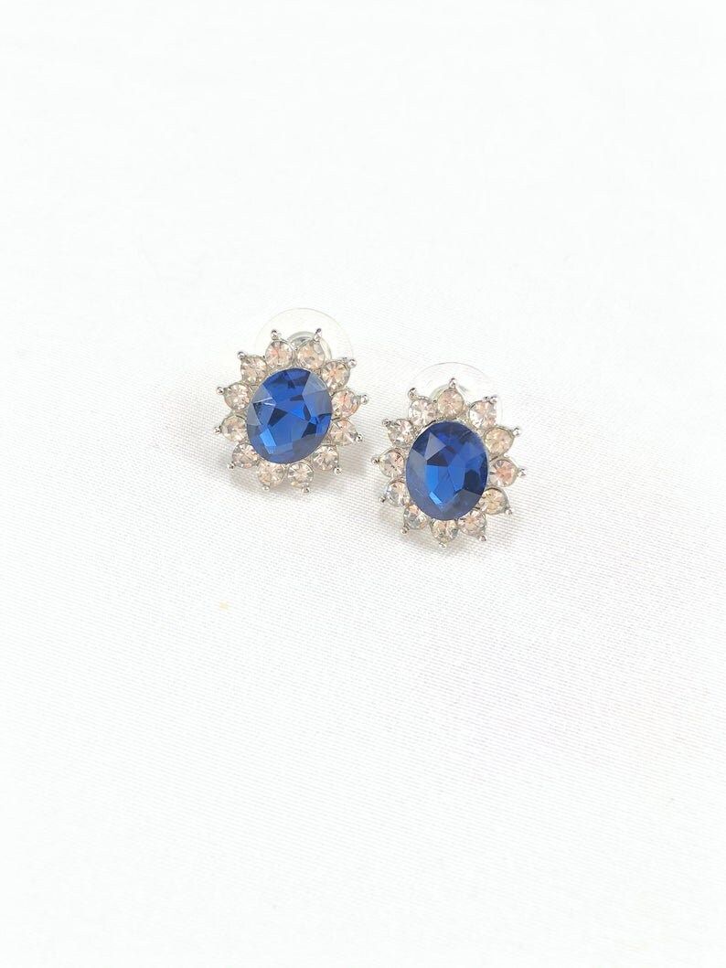 May include: A pair of silver stud earrings with a large blue oval-shaped gemstone surrounded by smaller clear gemstones.