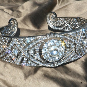 Puede incluir: Una diadema plateada con un dise&ntilde;o geom&eacute;trico y un gran strass transparente en el centro. La diadema est&aacute; decorada con strass m&aacute;s peque&ntilde;os.