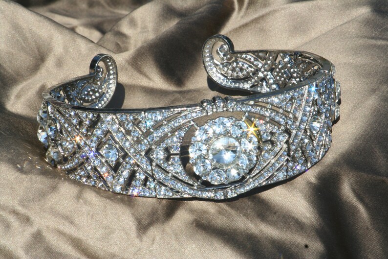 Puede incluir: Una diadema plateada con un dise&ntilde;o geom&eacute;trico y un gran strass transparente en el centro. La diadema est&aacute; decorada con strass m&aacute;s peque&ntilde;os.