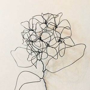 May include: A black wire sculpture depicting a flower, likely a hydrangea, set against a plain backdrop. The piece showcases intricate petal and leaf details, complete with a stem and a hanging hook. The design is modern and minimalist.