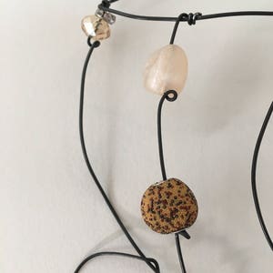 May include: A black wire necklace with three beads. The beads are a light brown with a speckled pattern, a light pink, and a clear bead with a silver accent.