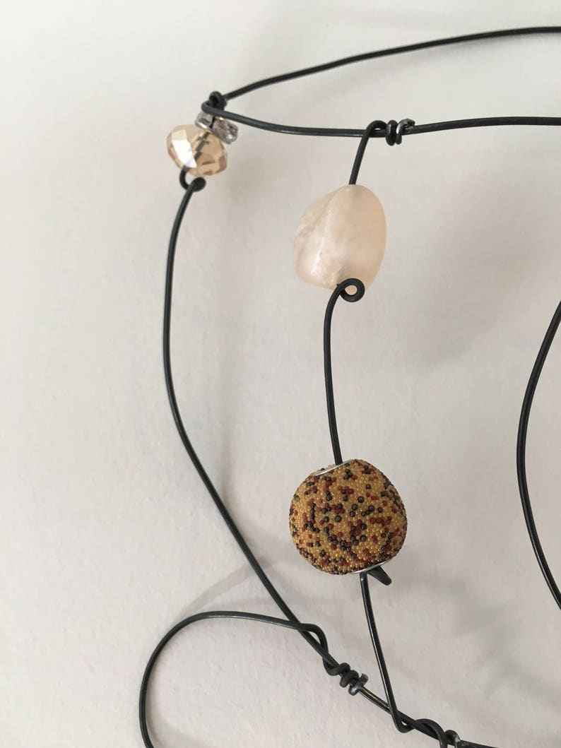 May include: A black wire necklace with three beads. The beads are a light brown with a speckled pattern, a light pink, and a clear bead with a silver accent.