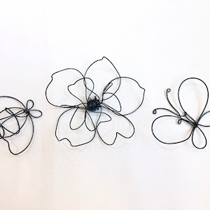 May include: Three wire sculptures: a flower, a butterfly, and a smaller butterfly. The flower is made of wire and has a simple, elegant design. The butterflies are also made of wire and have delicate wings.