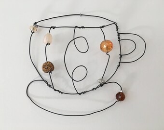 May include: A wire sculpture of a coffee cup with a saucer, decorated with brown, beige, and orange beads.