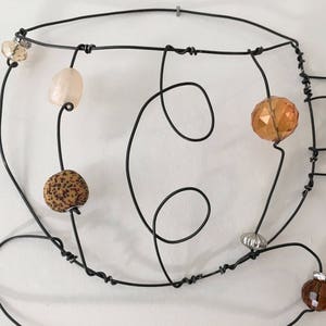 May include: A wire sculpture of a coffee cup with various colored beads and glass accents. The sculpture is made of black wire and features a brown, beige, and orange bead.