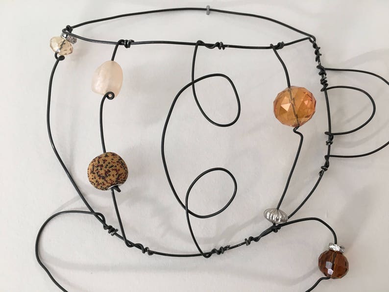 May include: A wire sculpture of a coffee cup with various colored beads and glass accents. The sculpture is made of black wire and features a brown, beige, and orange bead.