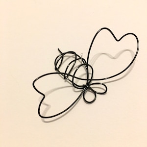 May include: A black wire sculpture in the shape of a butterfly. The sculpture has two heart-shaped wings and a tangled body. A small metal ring is attached to the body. The sculpture is on a white background.