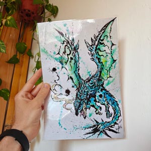 Può includere: Stampa artistica fantasy di un drago con dettagli acquerellati blu e verde acqua. Il drago è raffigurato in volo con le ali spiegate, su sfondo bianco con schizzi di vernice verde acqua e nera. L'opera d'arte è racchiusa in una custodia protettiva trasparente.