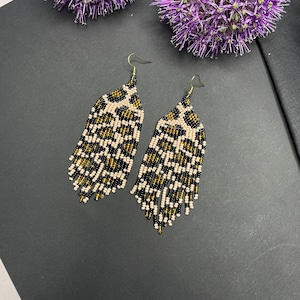 May include: A pair of beaded earrings with a leopard print design. The earrings feature a cream, black, and gold colour scheme. The earrings have a teardrop shape with a fringe at the bottom and gold-coloured hooks.