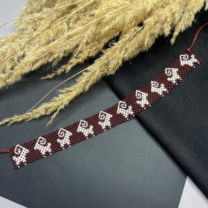 May include: A brown beaded bracelet with a white geometric pattern. The bracelet is made of small beads and has a simple design.