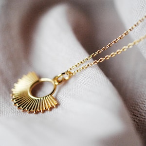 Könnte beinhalten: Eine goldfarbene Halskette mit einem Sonnenstrahl-Anhänger. Der Anhänger hat ein kreisförmiges Design mit radialen Linien, das an einer zarten Kette hängt. Die Halskette wird auf einem weichen, weißen Stoff präsentiert.
