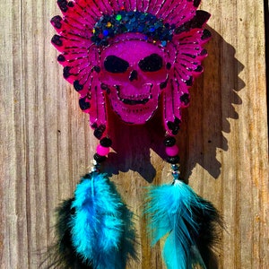 May include: A pink skull wearing a pink and black headdress with glitter and blue and black feathers hanging from the bottom.