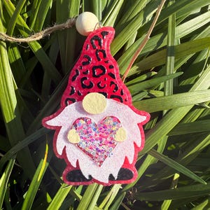 May include: A gnome-shaped ornament with a red leopard print hat and a white beard. The gnome holds a heart filled with glitter. A wooden bead and twine are attached for hanging. The ornament is set against a backdrop of green foliage.