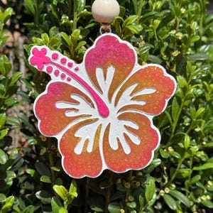 May include: A pink and orange glitter hibiscus flower ornament with a white center. The flower is hanging from a string with a wooden bead.
