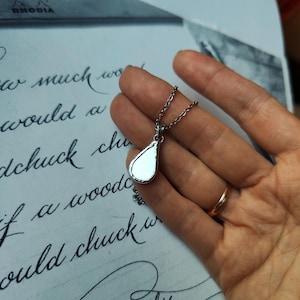 Puede incluir: Un collar de plata con un colgante en forma de lágrima y una cadena. El colgante es liso y reflectante.